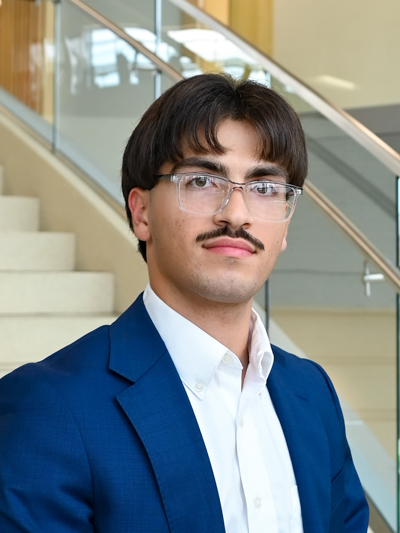 Man in glasses and blue blazer standing by a staircase indoors.