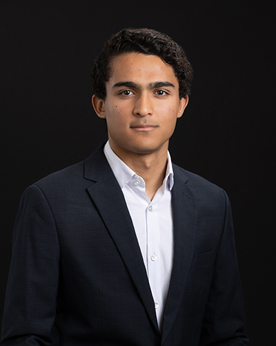 Young man in a dark suit and white shirt, facing forward against a black background.