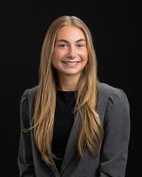 Smiling person with long blonde hair, wearing a gray blazer and black shirt against a black background.