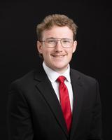 Man in glasses wearing a black suit, white shirt, and red tie, smiling against a black background.