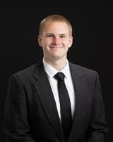 Man in a black suit and tie smiling against a dark background.