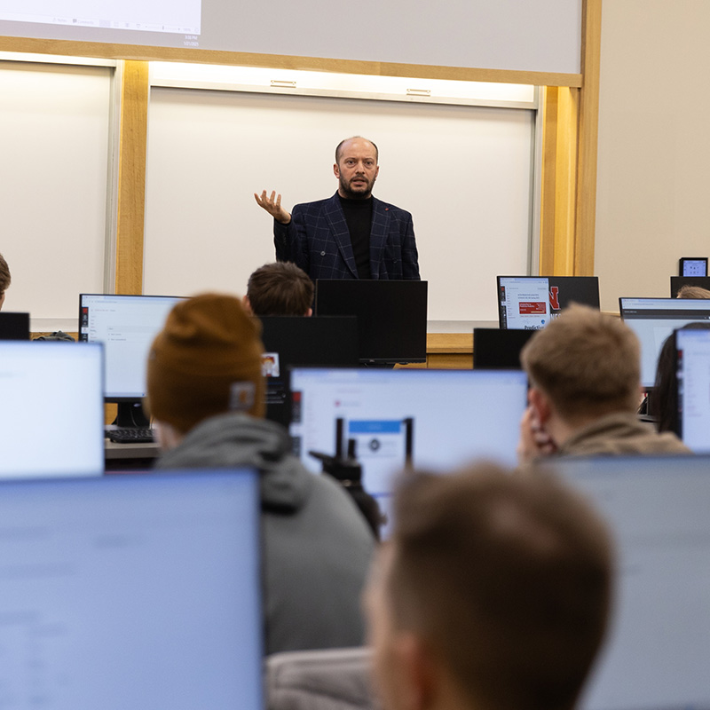 A lecturer gestures in a classroom with students using computers.