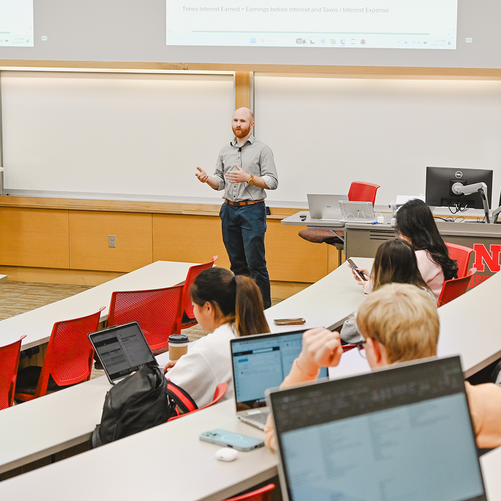 Man speaking to students in a classroom with a presentation screen in the background.