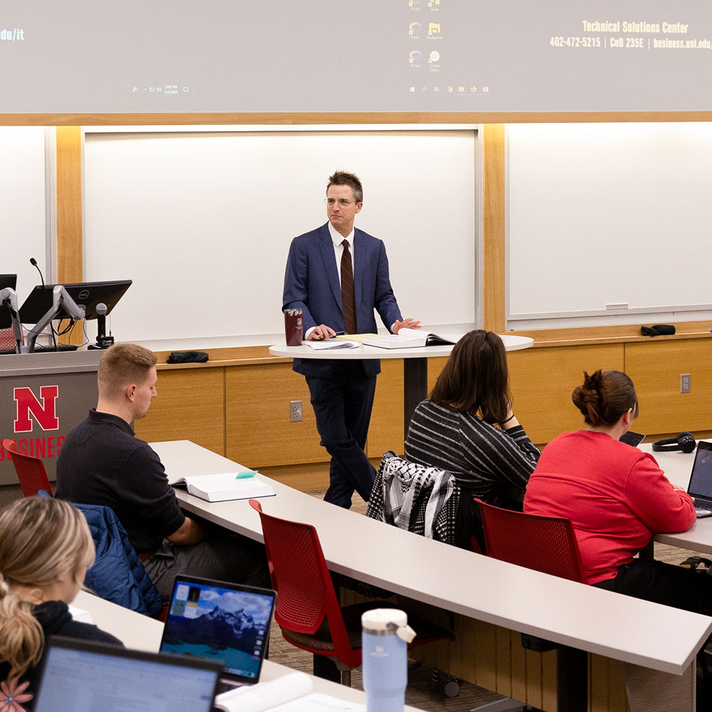 Speaker in formal attire addressing a classroom of seated attendees under a bright screen.