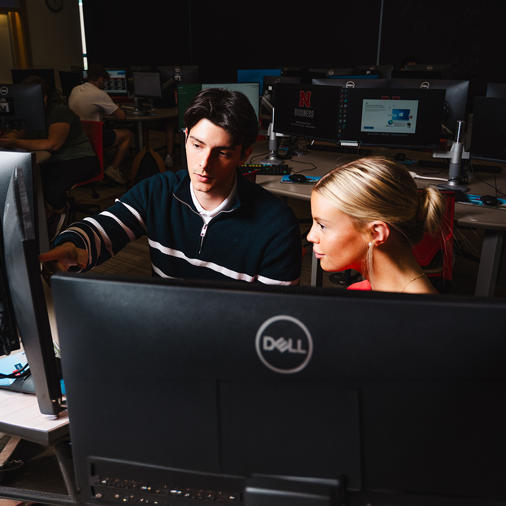Two people collaborating at a computer desk in a dimly lit classroom with multiple monitors.