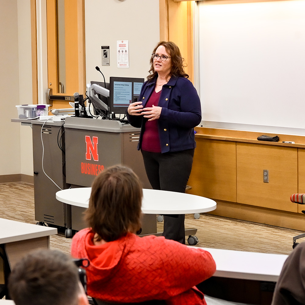A speaker gestures during a presentation in a classroom with attendees seated in the foreground.