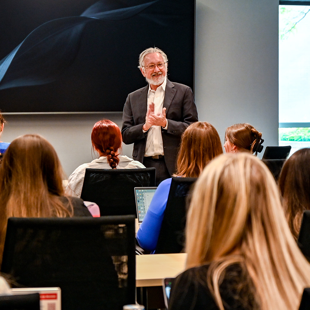 A man speaking to seated attendees in a conference room with a large screen behind him.