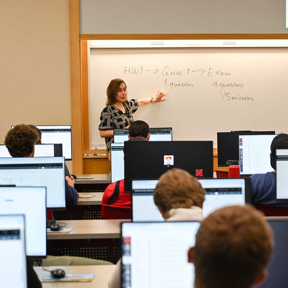 Teacher pointing at a whiteboard in a computer lab classroom; students seated at desks.