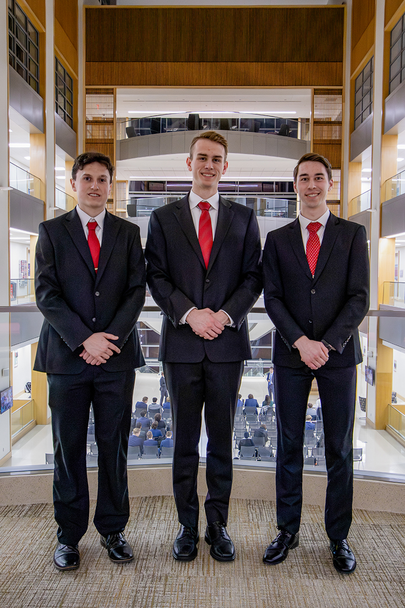Three men in black suits and red ties stand side by side in a modern, well-lit building.