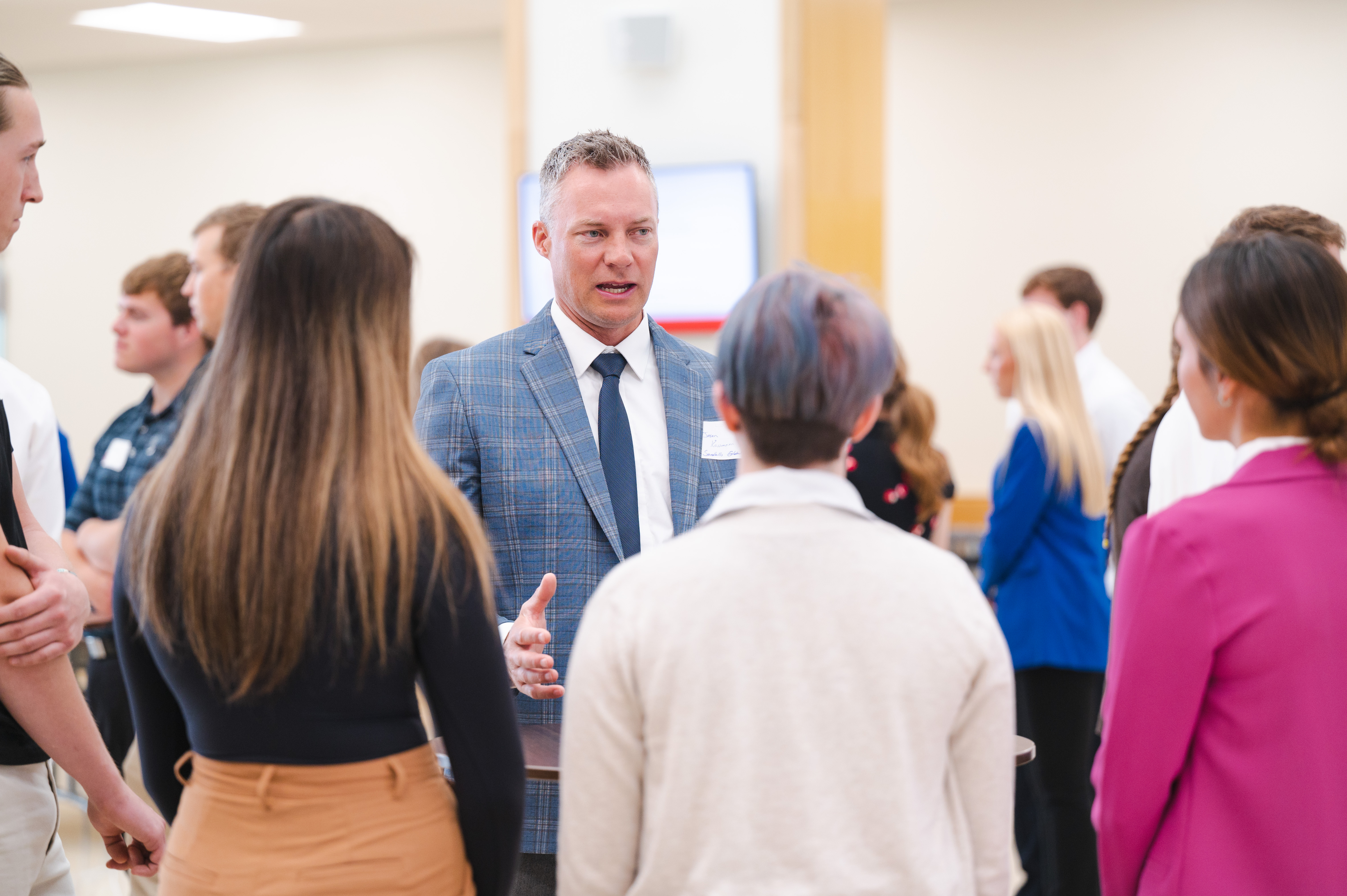 Man in a suit speaking to a group of people in a bright indoor setting.