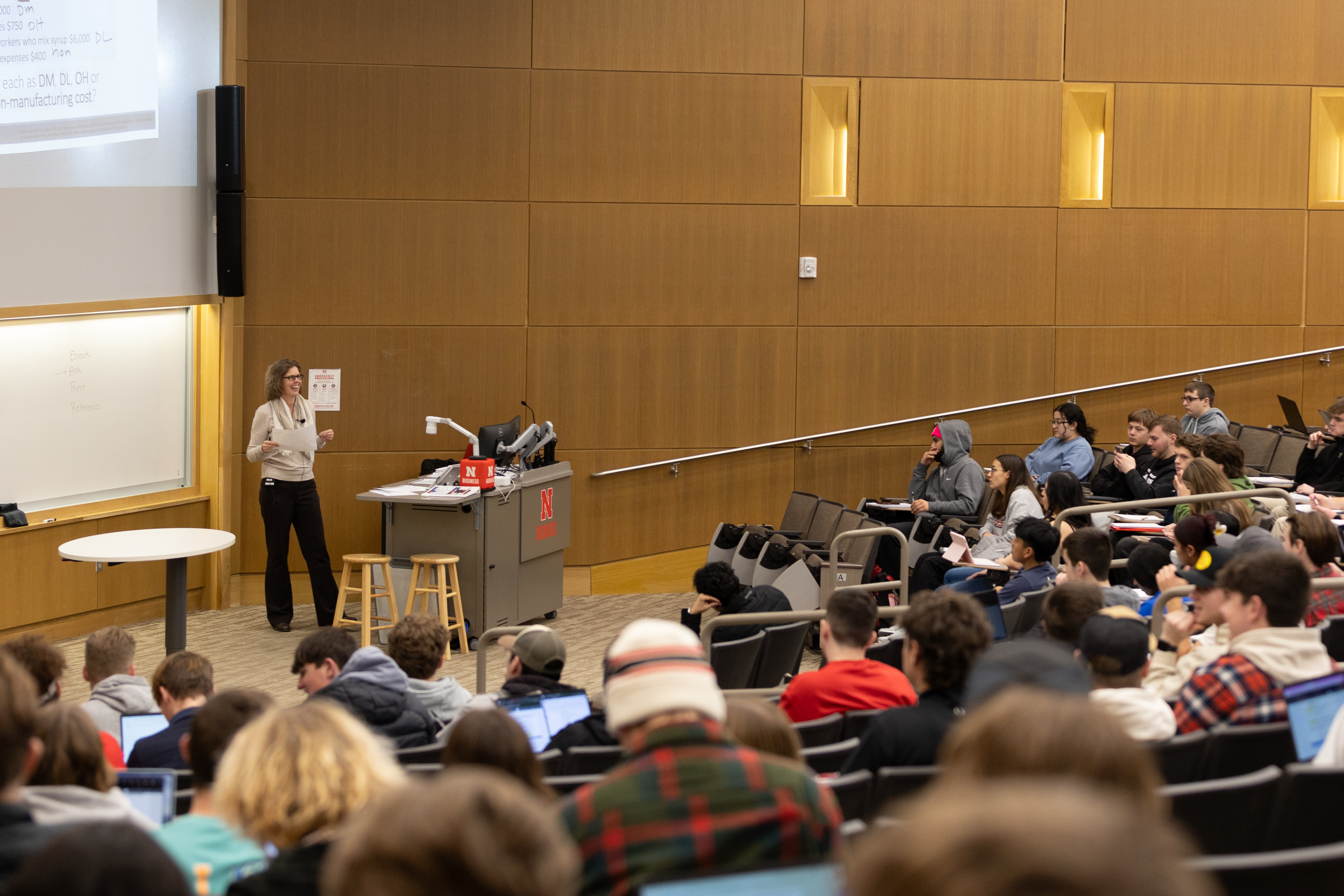 Lecture hall with a professor speaking and students seated, some using laptops.