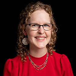 Curly-haired person wears a red blouse, beaded necklaces, and earrings, with a black background.