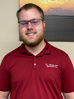 Smiling man in a red polo shirt stands indoors with a sunset photo hanging behind him.