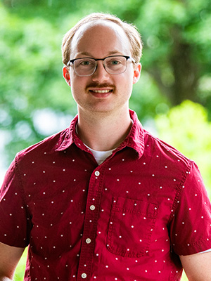 Man with glasses and a mustache wearing a red shirt, standing outdoors with greenery in the background.