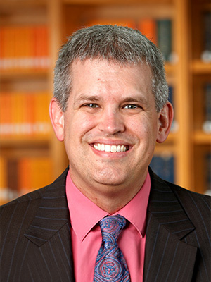 Man in a suit with a pink shirt and colorful tie, smiling in a library setting.