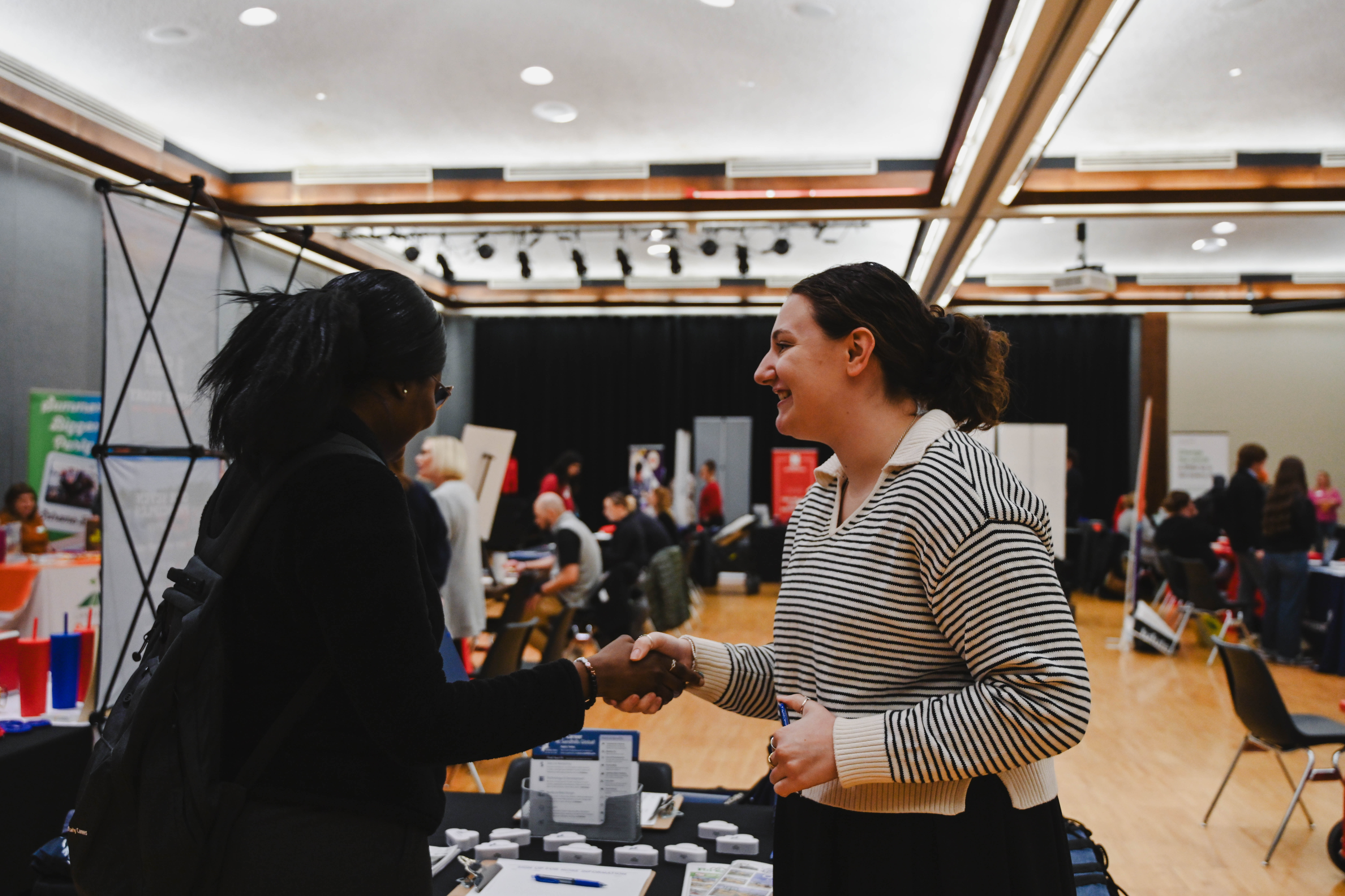 Two women shaking hands at a career fair in a bright, open room with tables and displays.