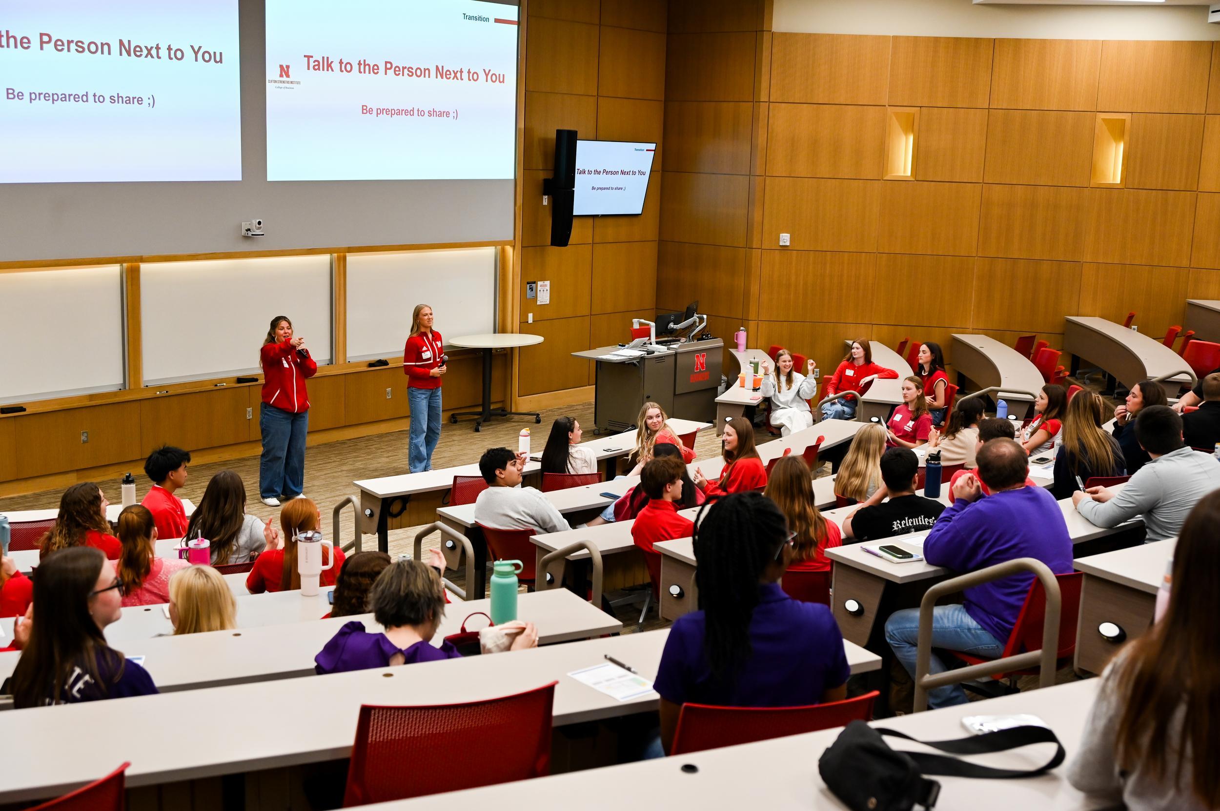 Lecture hall with two presenters and an audience of students seated in tiered rows.
