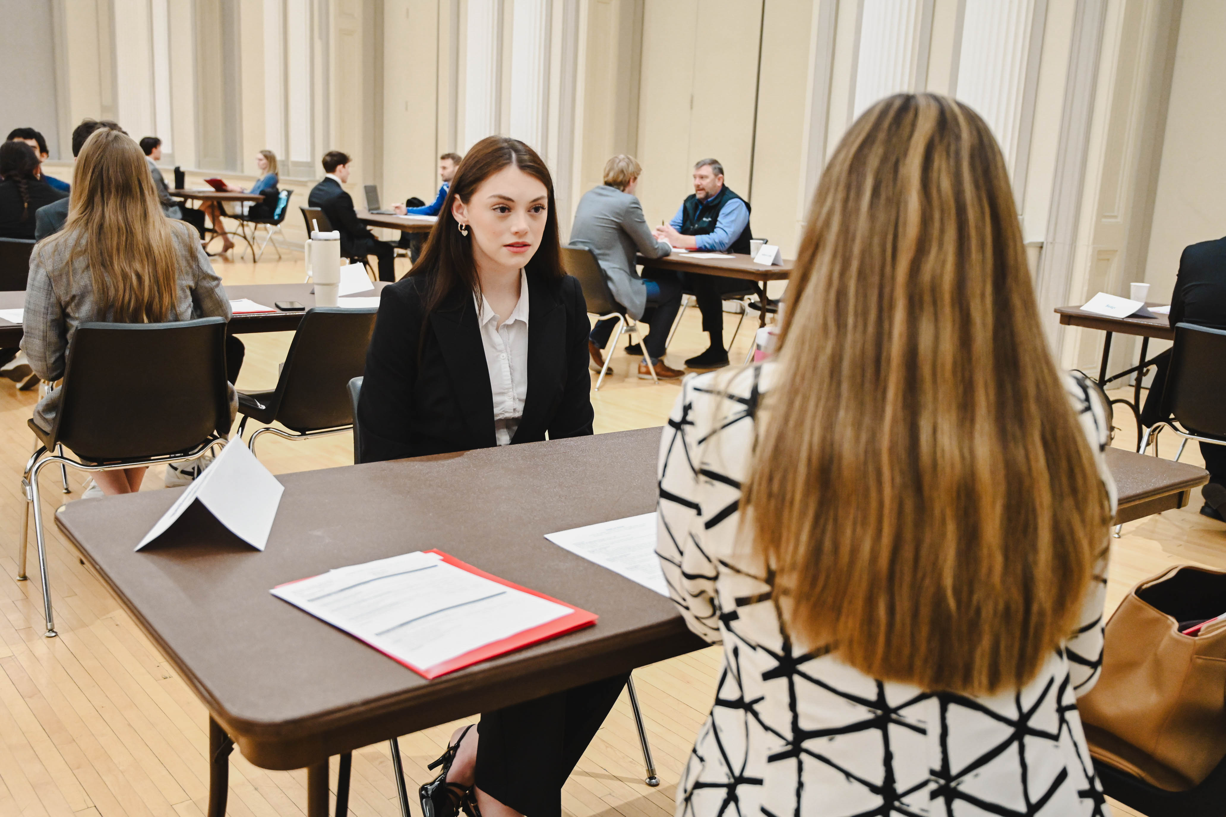 Two women seated at a table during a professional interview in a hall with others nearby.