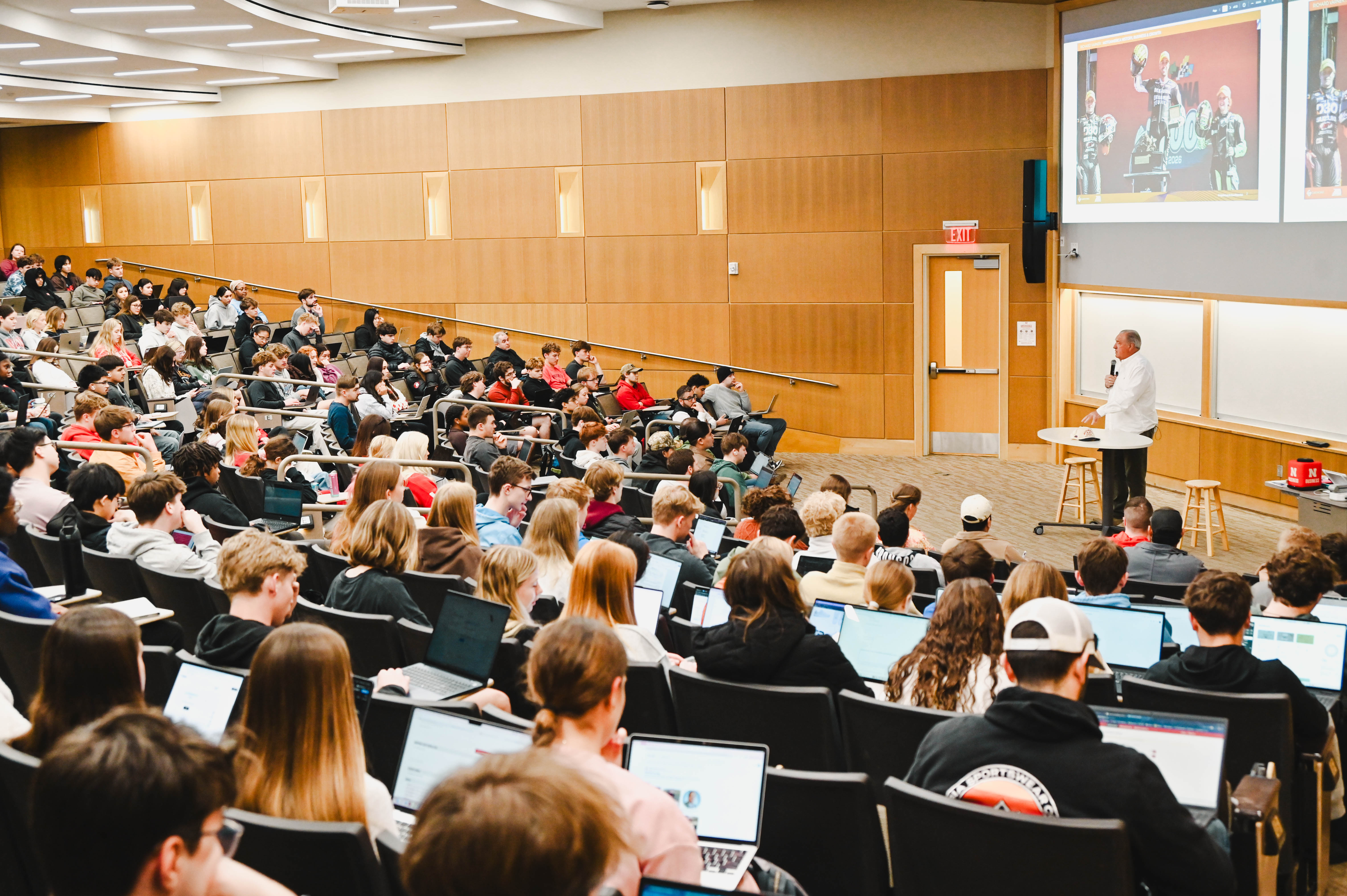 Large lecture hall with students seated, many using laptops, as a professor presents at the front.