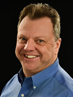 Smiling man in a blue shirt against a black background.
