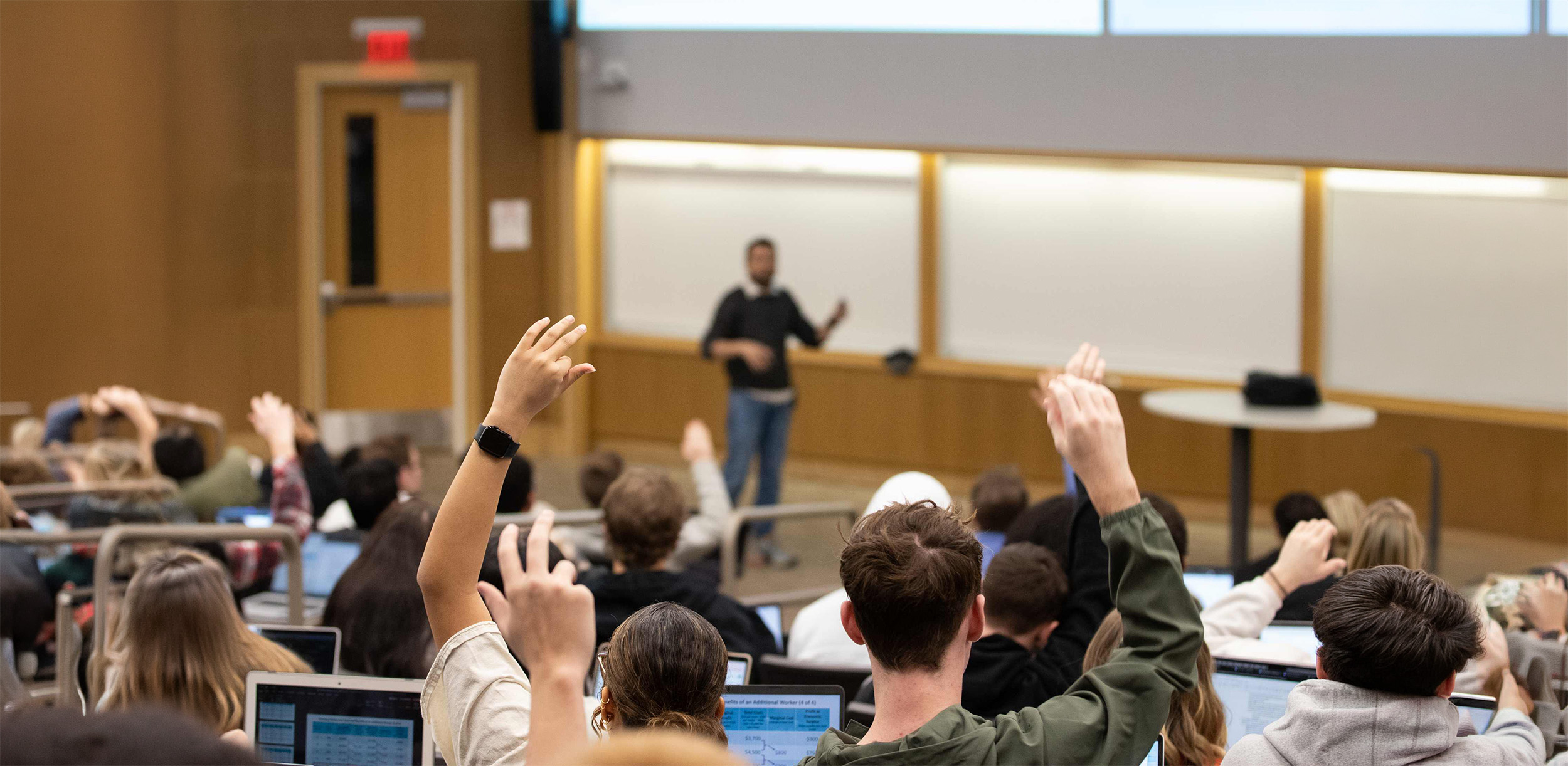Students raising hands in a lecture hall while a speaker presents at the front.