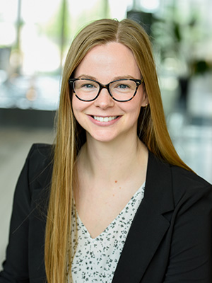Smiling woman with long blonde hair, black glasses, and a black blazer in a bright indoor setting.
