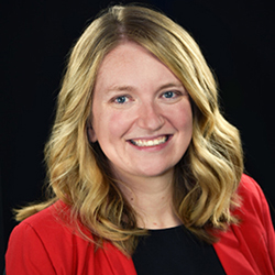 Woman wearing a red blazer, blonde wavy hair, blurred face, dark background.