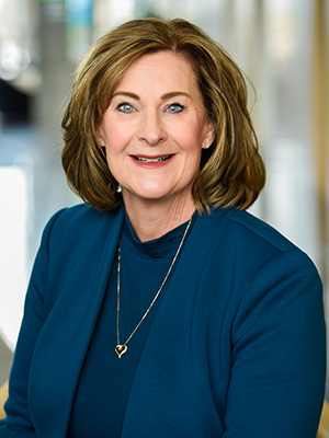 Smiling woman with shoulder-length hair wearing a blue blazer and heart-shaped necklace.