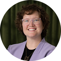 Smiling woman with curly hair, glasses, and a lavender blazer in front of a dark green curtain.