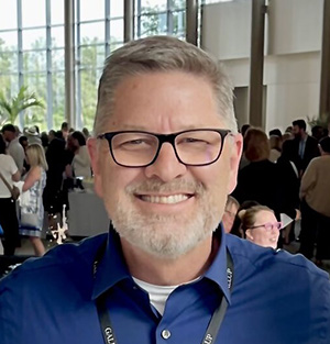 Man wearing glasses and a blue shirt, smiling in a brightly lit room with people in the background.