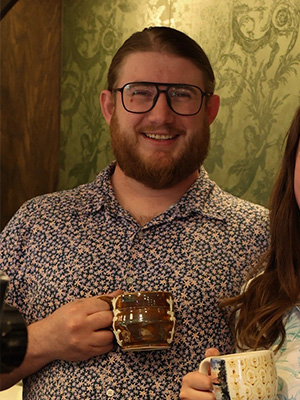 Man with glasses and a floral shirt smiling while holding a brown ceramic mug.