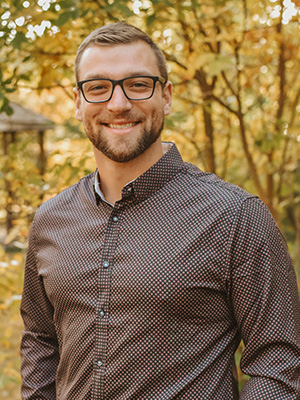 Smiling man wearing glasses and a patterned shirt, standing outdoors surrounded by autumn trees.