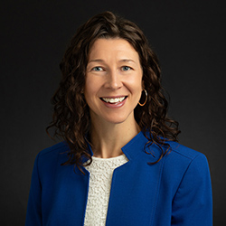 Dark-haired woman wearing a blue blazer and white top against a black background.