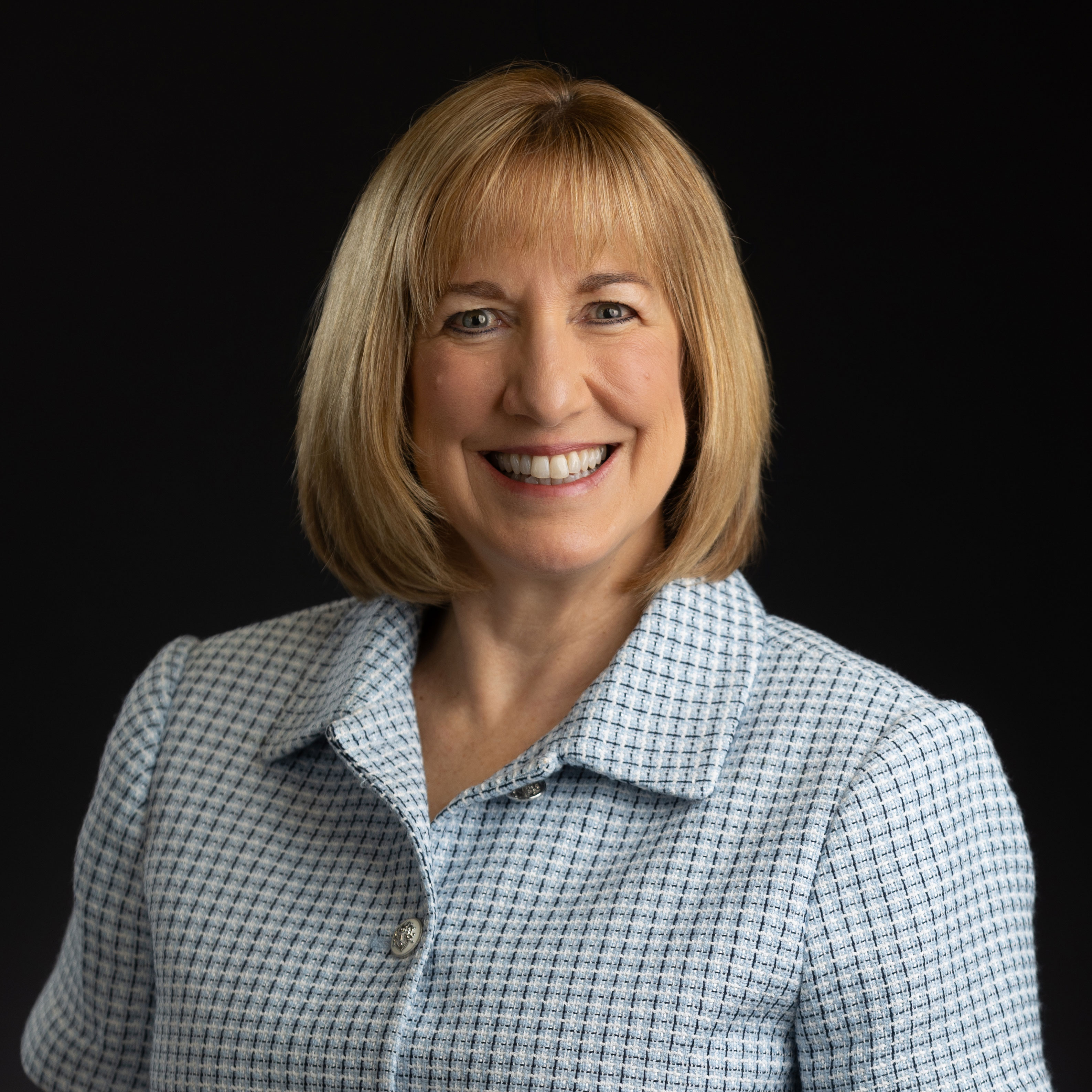 Smiling woman with blonde bob haircut in a checkered shirt against a black background.