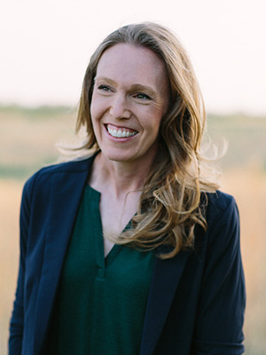 A smiling woman with long blonde hair wearing a dark blazer and green shirt, standing outdoors.