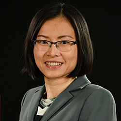 Woman with short black hair, glasses, and a gray blazer smiling against a black background.