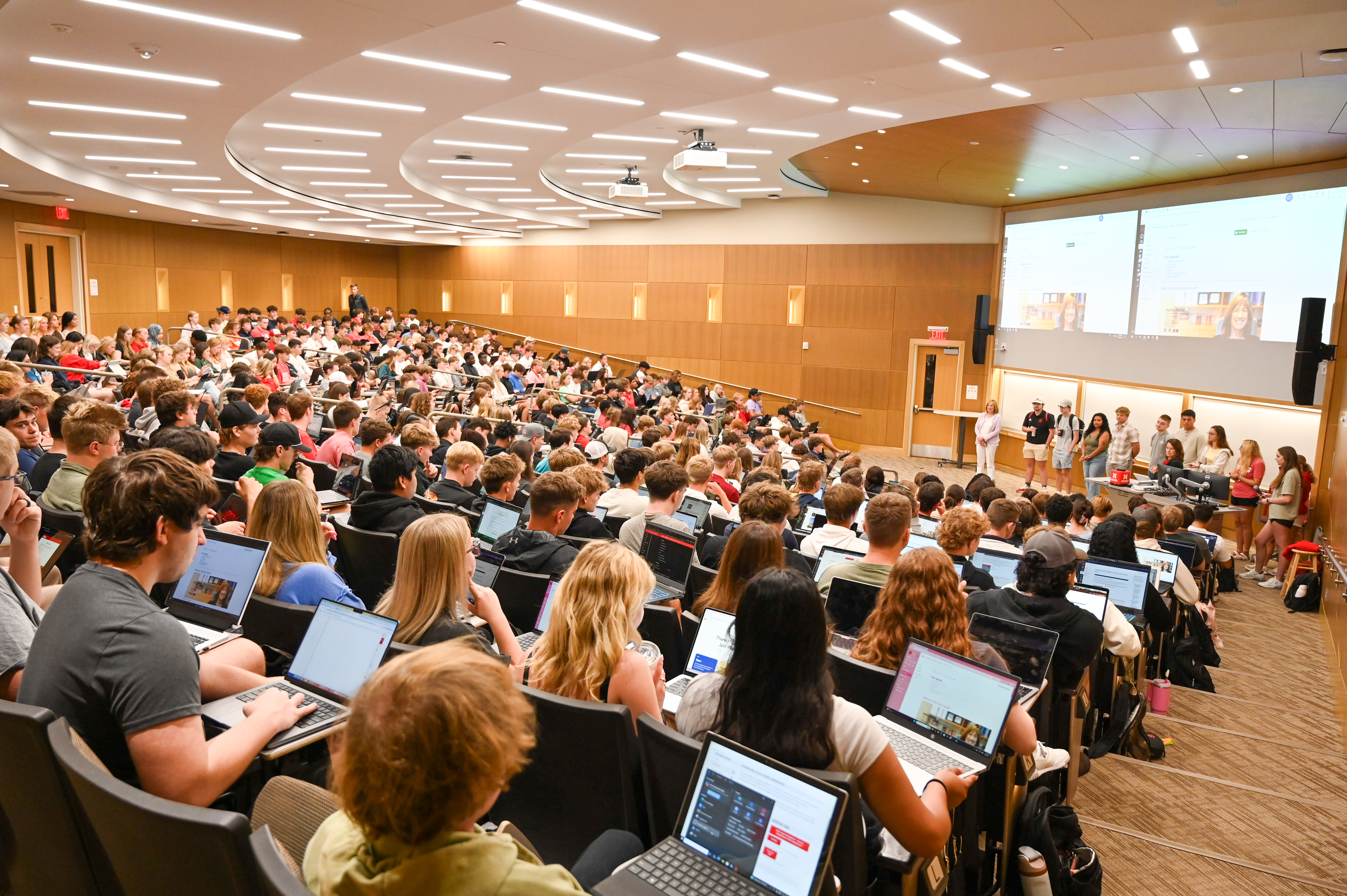 Large lecture hall filled with students, many using laptops; speakers present at the front.