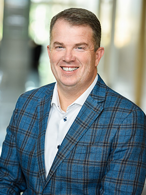 Smiling man in a blue plaid blazer and white shirt, standing indoors with blurred background.