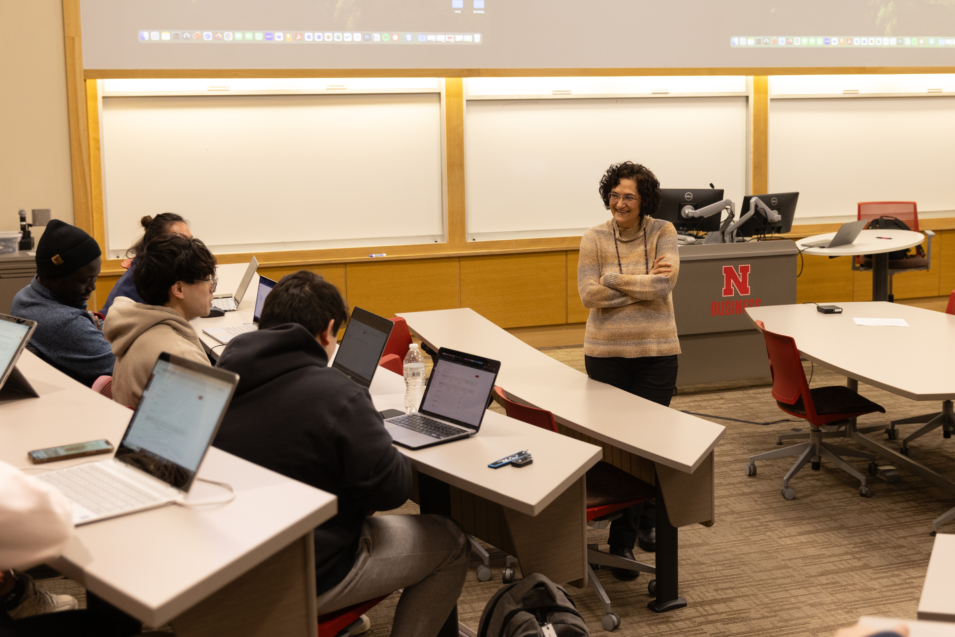 Classroom with students at desks and a presenter standing near the podium, projected screen above.