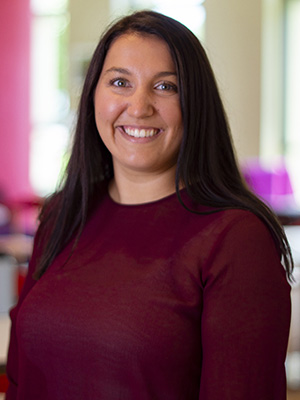 Smiling woman in a maroon top standing indoors with a blurred colorful background.