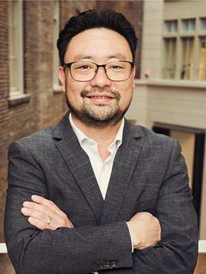 A man in a suit, smiling with arms crossed, standing in an urban setting with brick buildings.
