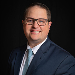Man in a dark suit and light tie against a black background. Face blurred.