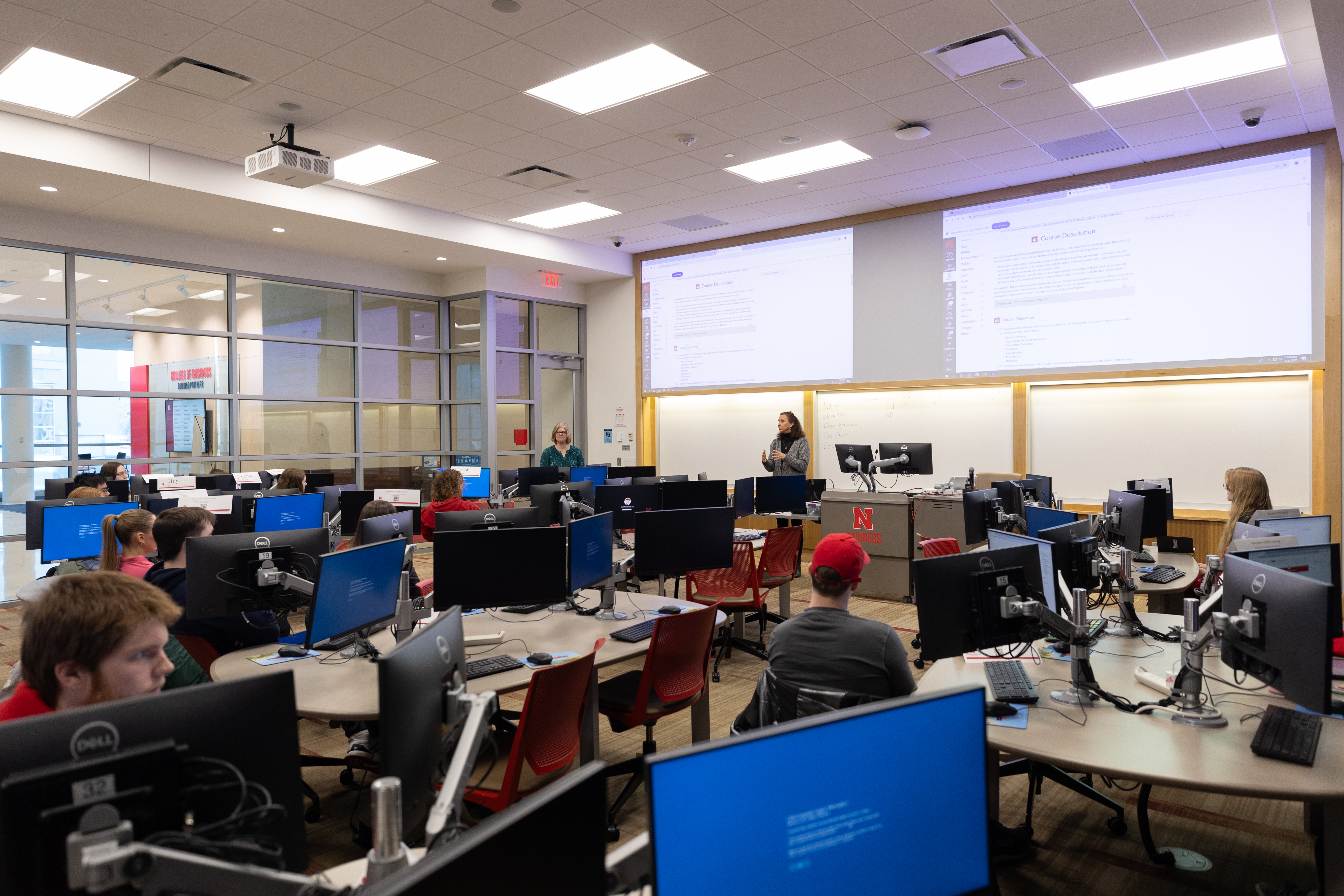Classroom with students seated at computer stations, instructor teaching at the front, screens displayed.