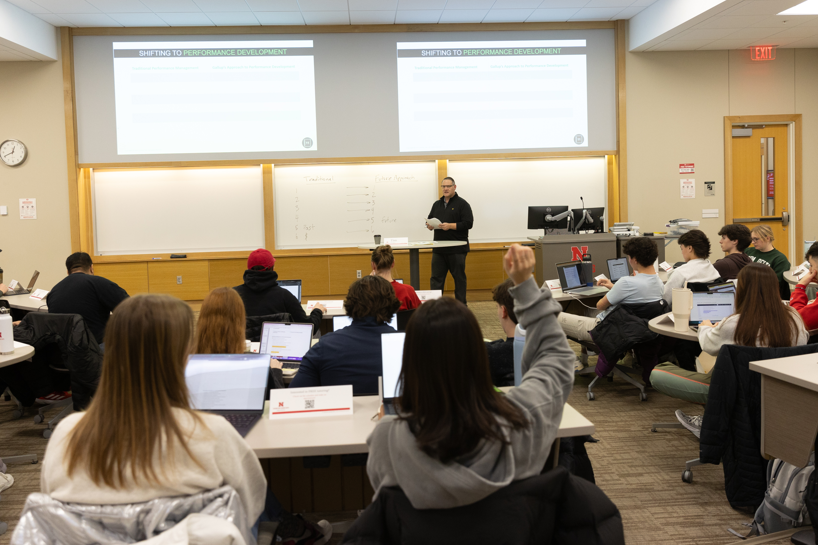 Classroom setting with a teacher speaking to students, one raising a hand, and others using laptops.