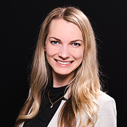 Blonde-haired person wearing a white blazer and black top against a dark background.