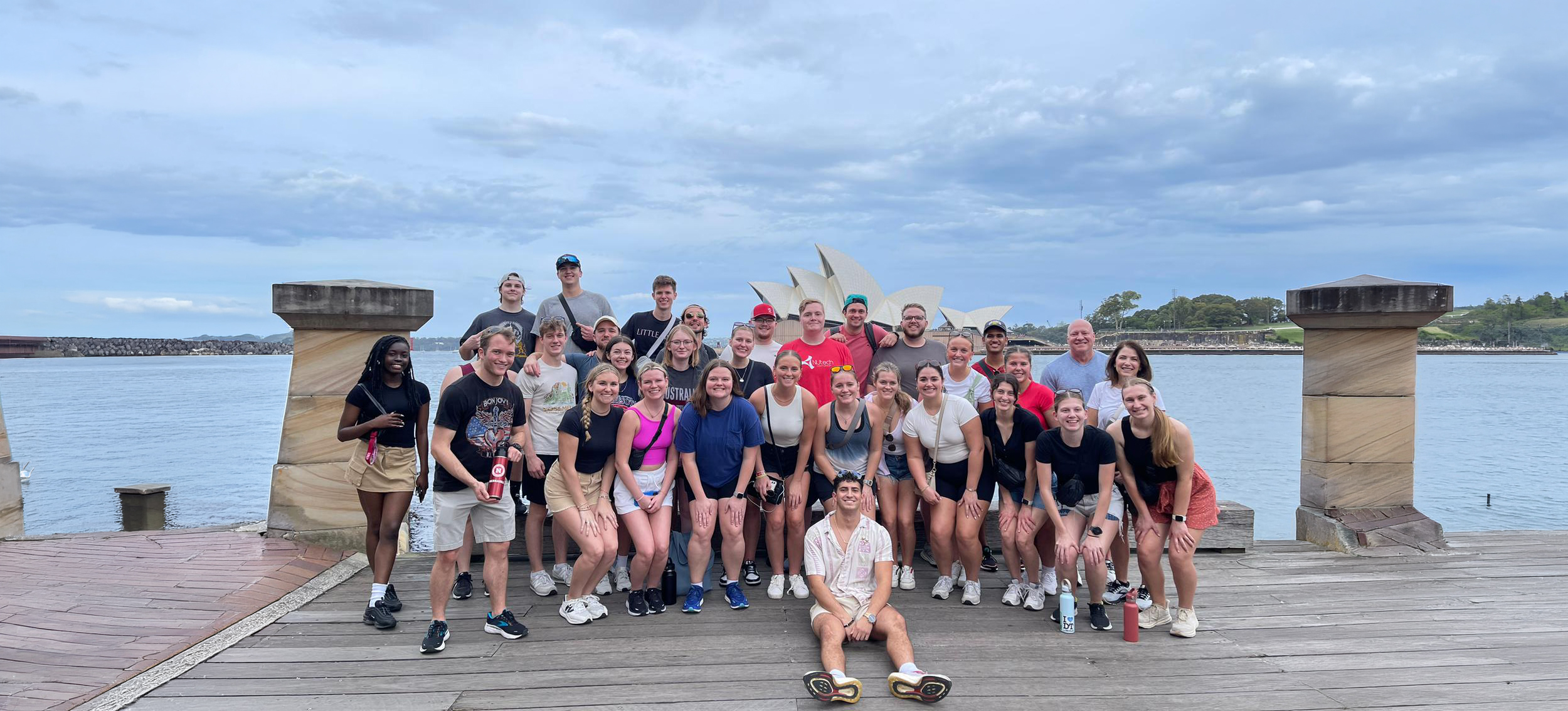 Group of people smiling on a wooden platform by water with the Sydney Opera House in the background.