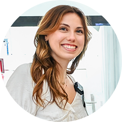 Smiling woman with wavy brown hair wearing a white shirt and a device on her shoulder.