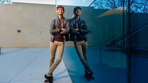 Aaryan Naik leans against Hawks Hall for a photo with his reflection in the glass of the building