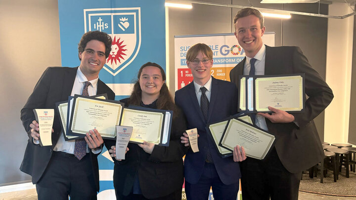 Group of four students in formal business attire holding certificates and trophies.