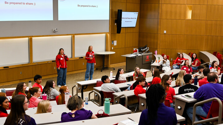 Classroom with students seated in rows and two presenters speaking at the front near a projector screen.